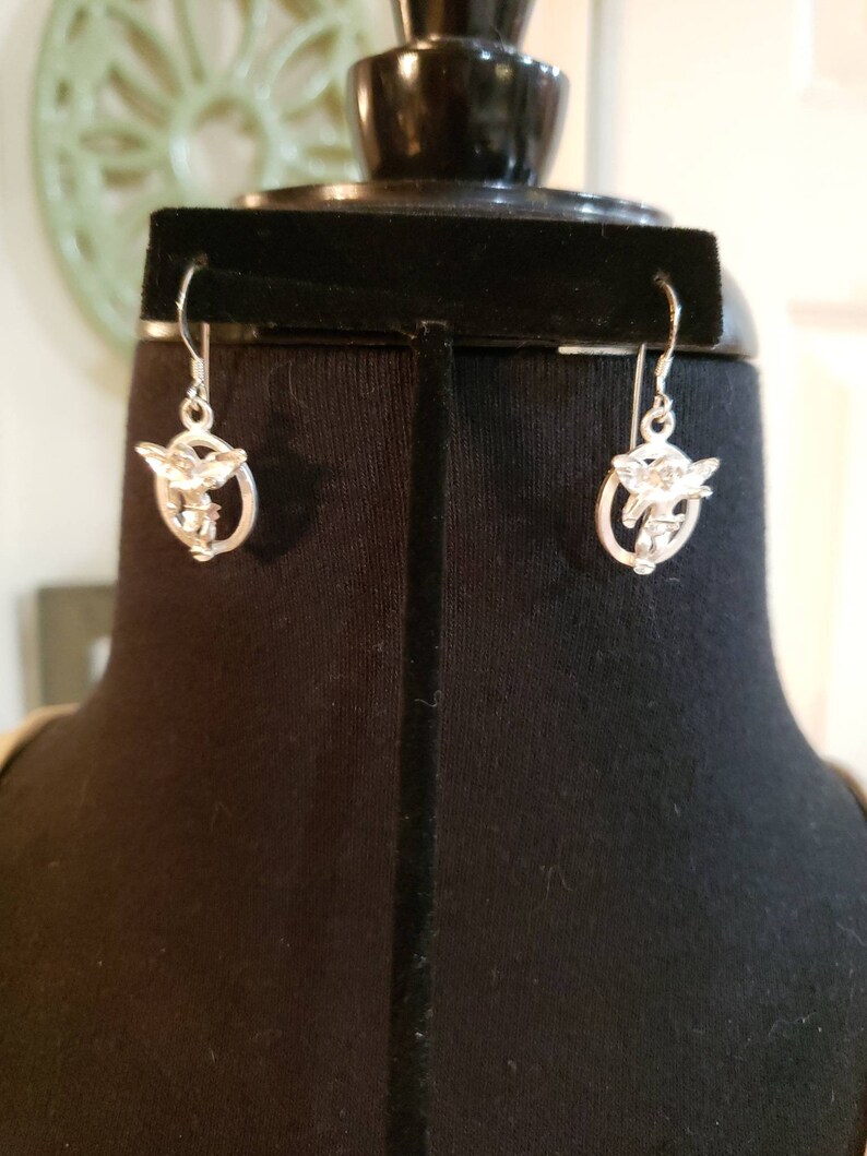 May include: A pair of silver dangle earrings featuring a cherub design. The cherubs are holding a heart and arrow.