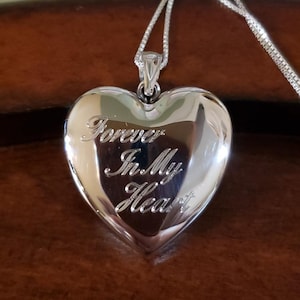 May include: Silver heart-shaped locket necklace with a chain. The locket has the inscription "Forever In My Heart" engraved on the front.