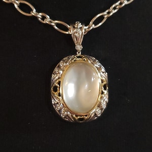 May include: A silver chain necklace with a pendant featuring a large oval-shaped white gemstone surrounded by a gold filigree design and small clear gemstones.
