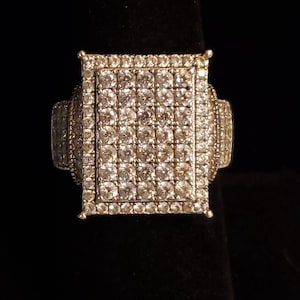 May include: A silver ring with a large square-shaped center stone set with many small clear stones.