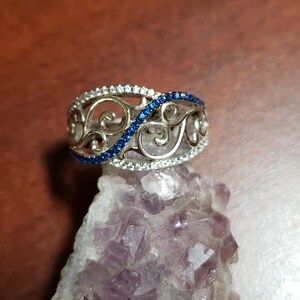 May include: A silver ring with a delicate filigree design and a row of blue gemstones. The ring is set on a purple amethyst crystal.