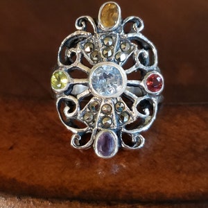 May include: A silver ring with a filigree design and a variety of gemstones, including amethyst, topaz, citrine, and garnet. The ring is set with a large, clear center stone.