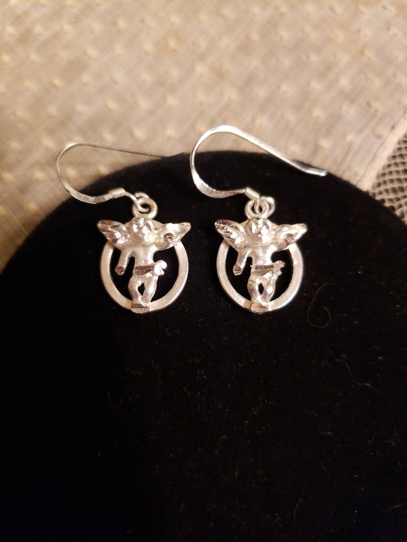 May include: A pair of silver earrings featuring a cherub design. Each earring has a small oval frame with a cherub figure inside.
