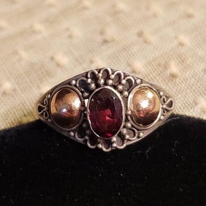 May include: A silver ring with a red gemstone and two gold accents. The ring has a delicate, ornate design.