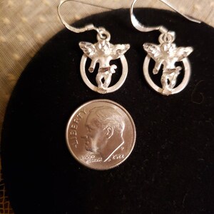 May include: A pair of silver earrings featuring a cherub design. Each earring has a small cherub figure with wings, standing in a circular frame.
