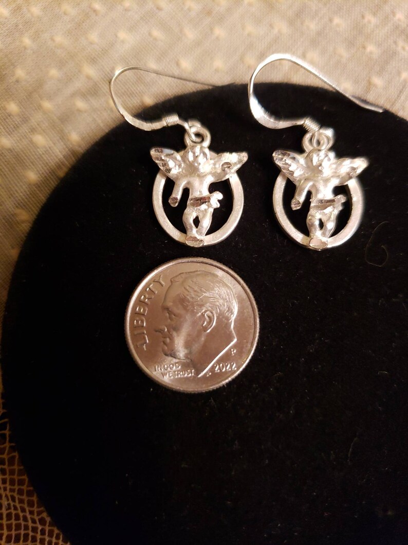 May include: A pair of silver earrings featuring a cherub design. Each earring has a small cherub figure with wings, standing in a circular frame.