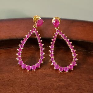 May include: A pair of gold dangle earrings with a teardrop shape. The earrings are adorned with pink gemstones.