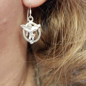 May include: Silver dangle earrings featuring a small angel charm with wings, inside a circular frame.