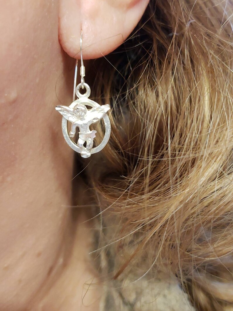 May include: Silver dangle earrings featuring a small angel charm with wings, inside a circular frame.