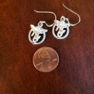 May include: A pair of silver earrings with a small angel design. The earrings are about the size of a US penny.