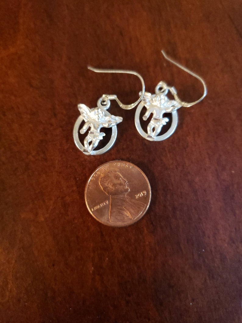 May include: A pair of silver earrings with a small angel design. The earrings are about the size of a US penny.