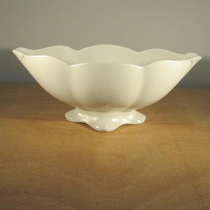 May include: Cream-coloured ceramic bowl with a wavy rim and a pedestal base. The bowl has a smooth, glossy finish and is placed on a wooden surface. The design is simple and elegant, suitable for serving or display.