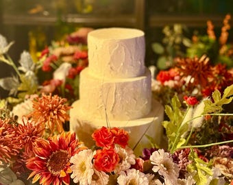 Puede incluir: Un pastel de boda blanco de tres pisos con un glaseado de crema de mantequilla texturizado, rodeado de un arreglo floral de flores naranjas, rojas y blancas.