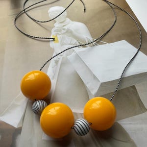 Puede incluir: Un collar llamativo con grandes cuentas esféricas amarillas brillantes, intercaladas con cuentas más pequeñas a rayas blancas y negras. Las cuentas están ensartadas en un cordón blanco y negro, creando un contraste llamativo.