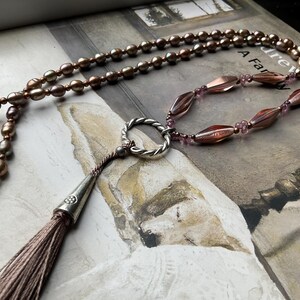 May include: A long necklace featuring a silver pendant and a tassel. The necklace is composed of brown and purple beads. The pendant is a circle with a small opening in the centre. The tassel is made of brown fabric.