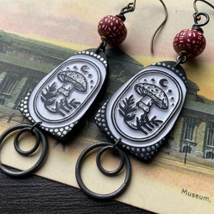 May include: A pair of black and white ceramic earrings with a mushroom and moon design. The earrings are attached to silver hoops with a red and white bead at the top.