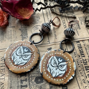 May include: A pair of owl earrings with a brown, white, and black color scheme. The owls are painted on a round, beige background with white polka dots. The earrings are attached to black metal hoops with a black and silver bead.