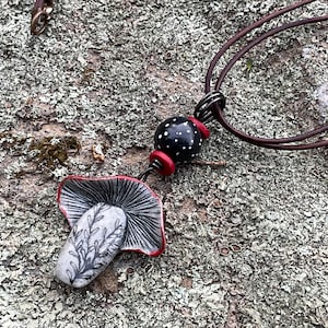 May include: A black and white ceramic mushroom pendant with a red rim hangs from a brown leather cord. The pendant is attached to a black and white polka dot bead with a red rim.