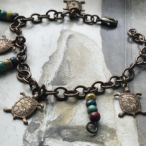Puede incluir: Una pulsera de cadena en tono bronce con abalorios de tortuga y cuentas de colores. La pulsera tiene un cierre de mosquetón y está adornada con múltiples colgantes de tortuga y pequeñas cuentas redondas de varios colores, creando un accesorio único.