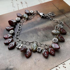 May include: A silver chain bracelet featuring leaf-shaped beads in shades of burgundy and olive green, accented with white leaf vein details. Small silver flower charms are interspersed throughout the design, creating a nature-inspired aesthetic.