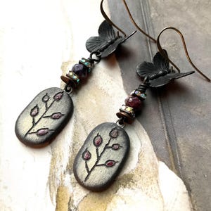 May include: A pair of handcrafted earrings featuring a butterfly accent and an oval pendant with a botanical design. The earrings have a dark metal finish, with red and iridescent bead accents. The oval pendants have a light gray background with a dark gray botanical design.