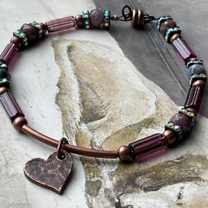 May include: A handmade bracelet featuring a copper-colored metal band and a heart-shaped charm. The bracelet is adorned with dark purple and clear glass beads, accented with iridescent rhinestones. The beads are a mix of cylindrical and faceted shapes.