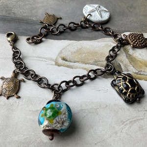 Könnte beinhalten: Ein bronzefarbenes Charm-Armband mit Schildkröten- und Seestern-Anhängern. Das Armband enthält eine blau-weiße Glasperle mit einem Schildkröten-Design. Die Kettenglieder sind oval.