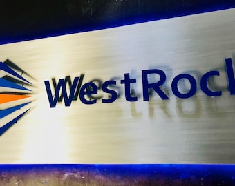 May include: A silver metal sign with the text "WestRock" in blue letters. The sign has a blue and orange geometric design in the lower left corner.