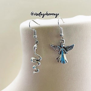 May include: A pair of silver earrings with a snake and angel design. The snake is coiled and the angel has wings outstretched. The earrings are hanging from a mannequin's neck.
