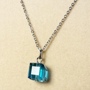 以下が含まれることがあります： 銀色のチェーンネックレスに青いクリスタル製のペンダントがついたもの。ペンダントは立方体で、ファセットカットが施されています。
