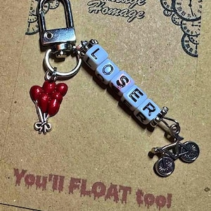 Könnte beinhalten: Ein silberner Schlüsselanhänger mit Karabinerverschluss, mit einem Anhänger aus roten Luftballons und einem Fahrrad. Das Wort "LOSER" ist mit weißen Buchstabenperlen geschrieben. Der Text "You'll FLOAT too!" ist auf der Karte gedruckt.