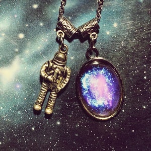 Puede incluir: Un collar de cadena de plata con dos colgantes. Un colgante es una figura de astronauta de plata. El otro colgante es un colgante redondo de vidrio con una imagen de galaxia púrpura y azul.