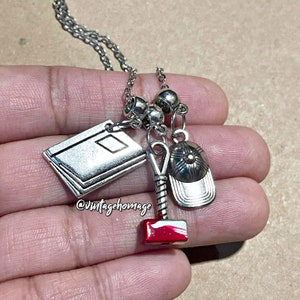 May include: Silver necklace with multiple charms, including a book, a baseball cap, a hammer, and a red gemstone. The necklace is on a hand.