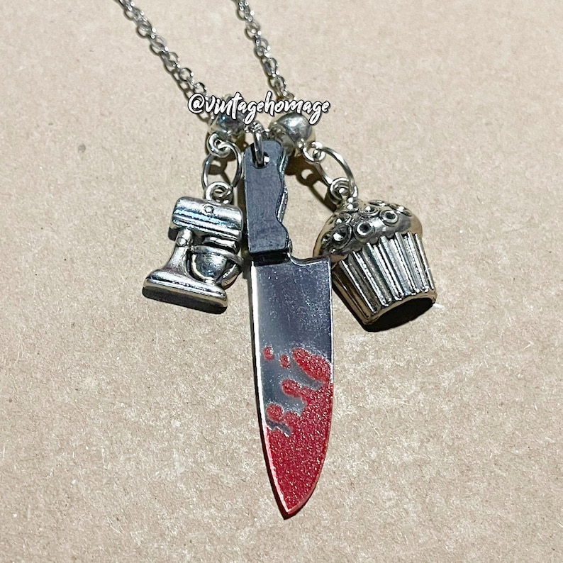 May include: A silver necklace with a bloody knife charm, a cupcake charm, and a miniature stand mixer charm. The necklace is on a brown surface. @vintagehomage