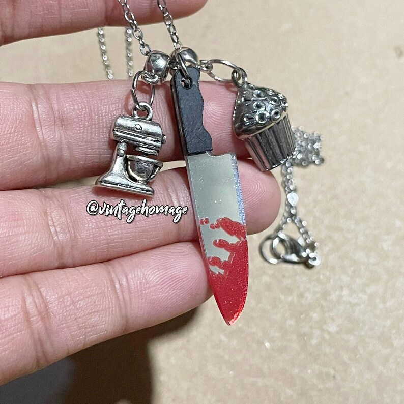 May include: A silver chain necklace with three charms: a silver cupcake, a silver kitchen mixer, and a silver knife with red paint on the blade. The necklace is held in a hand.