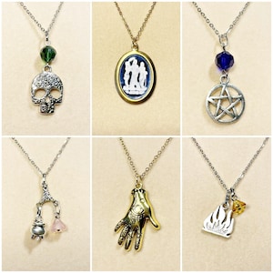 May include: A selection of silver-coloured necklaces, each with a unique pendant. Pendants include a skull, cameo, pentagram, cauldron, a golden hand with an eye, and a flame with a gemstone. Each necklace features a fine chain and colourful bead details.