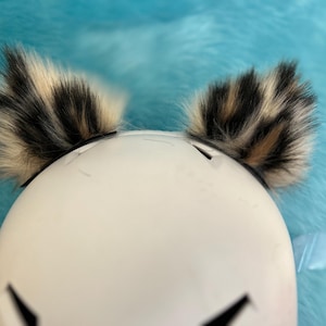 Könnte beinhalten: Weißer Helm mit schwarzen Akzenten und Kunstfellohren in Schwarz, Beige und Creme. Die Ohren sind an einem schwarzen Stirnband befestigt. Der Hintergrund ist eine hellblaue, flauschige Oberfläche.