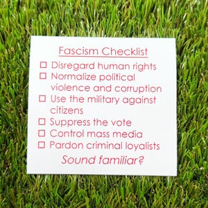 May include: A white card with the text "Fascism Checklist" in red font, listing actions such as disregarding human rights and suppressing the vote. The card is on a bed of green artificial grass. The text "Sound familiar?" is at the bottom.