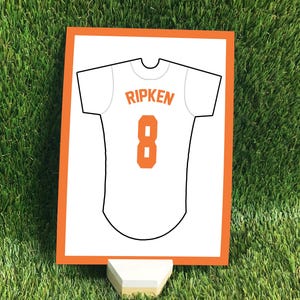 May include: A rectangular sign with an orange border featuring a white baseball jersey graphic. The jersey displays the name RIPKEN in orange letters above the number 8 in orange. The sign is supported by a small, light-colored wooden stand.