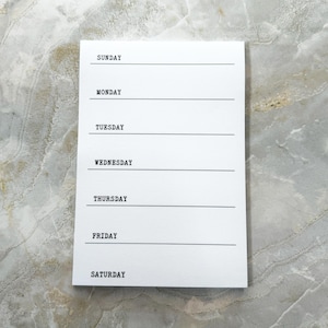 May include: A white rectangular weekly planner notepad featuring black typewriter-style text for each day of the week from Sunday to Saturday, with a horizontal black line under each day name to provide space for writing daily notes or tasks.