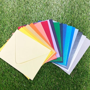 May include: A collection of colorful envelopes and matching paper fanned out on a bed of green grass. The envelopes and papers come in a variety of colors, including yellow, orange, red, and blue. The envelopes are square-shaped.