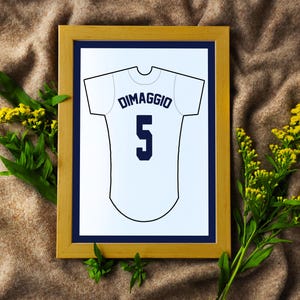 May include: A framed print featuring a white baseball jersey illustration with the name DIMAGGIO and the number 5 printed in dark blue. The artwork is displayed in a light wood frame with a dark blue mat, showcasing a clean graphic design.
