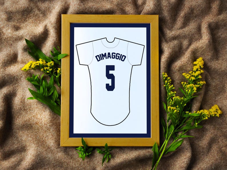 May include: A framed print featuring a white baseball jersey illustration with the name DIMAGGIO and the number 5 printed in dark blue. The artwork is displayed in a light wood frame with a dark blue mat, showcasing a clean graphic design.