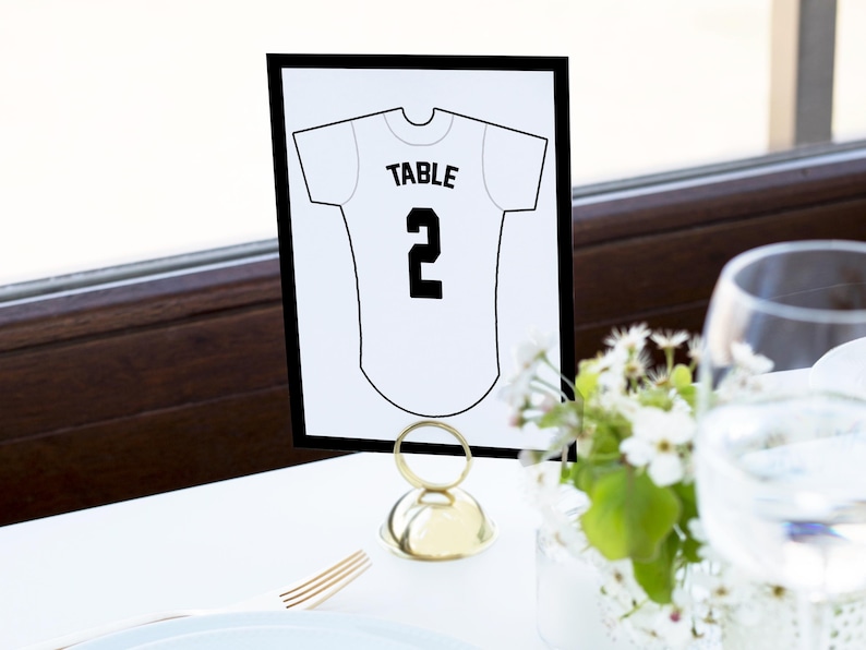 May include: A rectangular white table number card with a black border, featuring a black line drawing of a jersey with the text "TABLE" above the number "2" in the center. The card is held upright by a small, round, gold-colored metal stand.