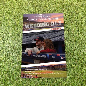 May include: A rectangular wedding day programme featuring a photograph of a couple at a baseball stadium. The text reads Jeffrey Cree Jr., Amber Nicole Martin, WEDDING DAY, PROGRAM, Saturday, April 30, 20XX, Fluor Field, 945 South Main Street, Greenville, SC 29601, 6:00 pm.