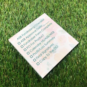 May include: A square paper checklist titled ASB Fundraising Checklist with a soft floral background. The list includes checkboxes for ASB Approval Date, Fundraiser Form Complete, PO For Product, Calendar HS/ASB/WEB, Profit/Loss Summary, Final Signatures, and Copy To The DO.