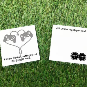 May include: Two white square cards on green grass. One card has a graphic of two gray game controllers connected by a heart-shaped cable, with the text "Life's better with you as my player two." The other card says "Will you be my player two?" and has two circles labeled "Player 1" and "Player 2."