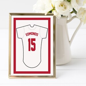 May include: A rectangular gold-toned frame containing a white print of a sports jersey. The jersey is outlined in black and features the name EDMONDS in red capital letters above the number 15 in red. A red rectangular border surrounds the jersey.