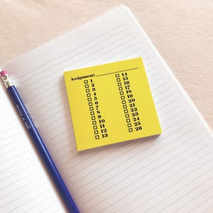 May include: A bright yellow square sticky note featuring the printed text Assignment: followed by two columns of numbered checkboxes from 1 to 26. The note is placed on an open notebook with lined pages next to a blue pencil with a pink eraser.