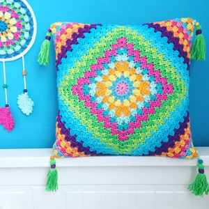 May include: A colourful, crocheted cushion with a geometric pattern in blue, green, pink, orange, and yellow. The cushion has green tassels at each corner. A dreamcatcher with similar colours hangs in the background.